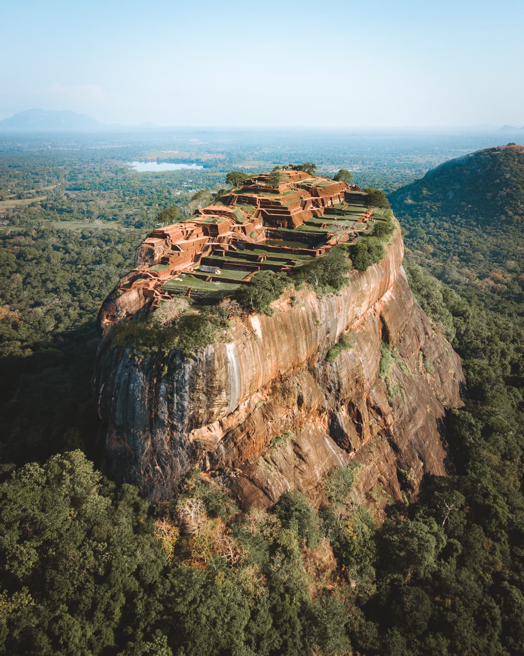 Aerial view of a flat-topped rock outcrop with ancient ruins on its summit, rising above surrounding forest.