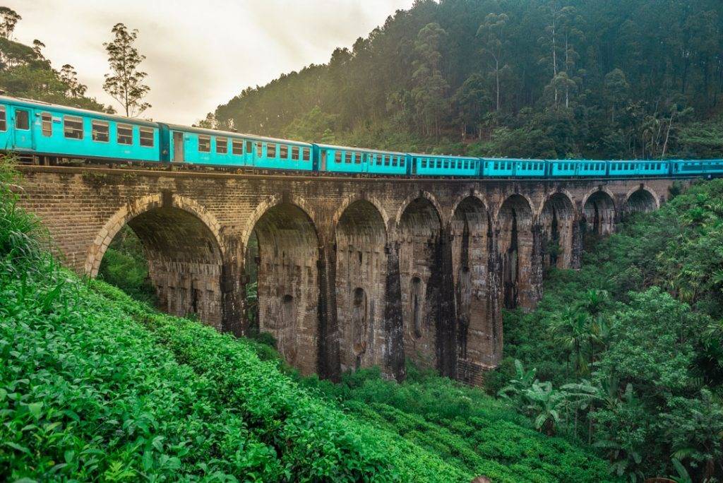 A turquoise train crossing a stone multi-arch railway viaduct, surrounded by forest.