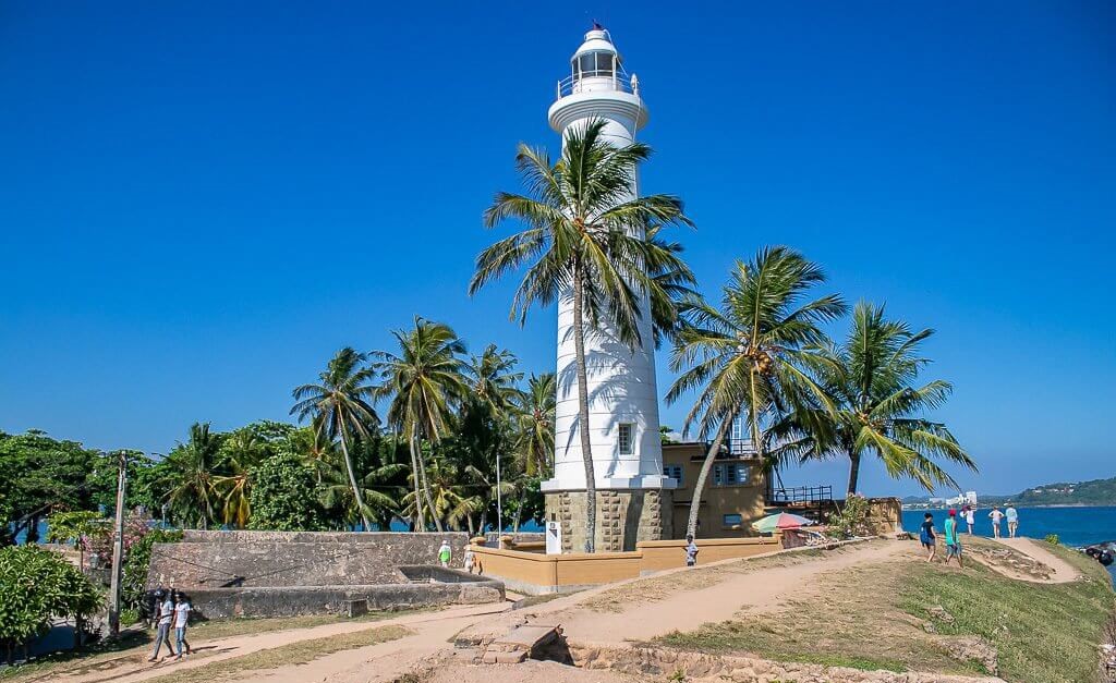 A white lighthouse surrounded by palm trees within a stone fort wall.
