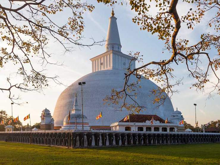A large white dome-shaped Buddhist stupa behind an elephant-relief retaining wall, decorated with prayer flags.