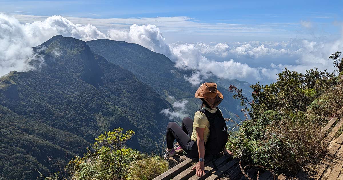 A hiker sitting on a cliff-edge staircase, looking out over cloud-covered mountains.