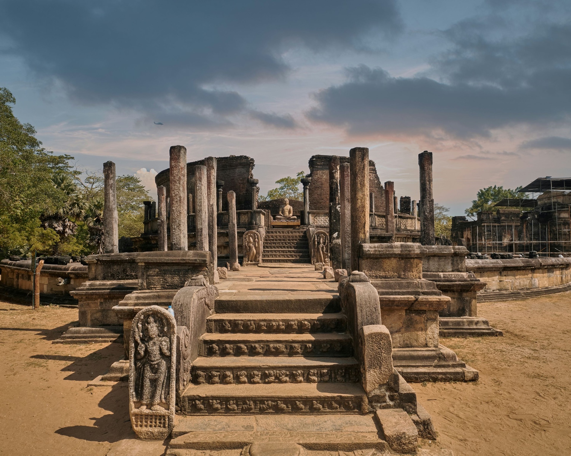 Ancient stone ruins with carved steps, guard stones and a seated Buddha statue among weathered columns.